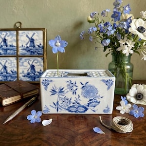 May include: A rectangular white ceramic flower frog vase featuring intricate blue floral patterns. The top surface has a grid of holes for individual stems, with one blue flower inserted. The design includes delicate botanical illustrations.