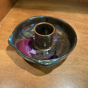 May include: A handmade ceramic candle holder with a dark, mottled glaze. The holder features a circular base with a raised center for the candle and a small spout. The color palette includes shades of brown, purple, and blue.