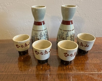 May include: Two ceramic sake carafes and four matching cups featuring a crackled cream glaze, a horizontal red band, and black Japanese calligraphy. The cups have a dark brown base and the text ひな鳥山 written in brown on the front.