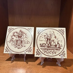 May include: Two square ceramic tiles with brown illustrations of Dutch scenes. One tile depicts a woman with a bucket, the other shows windmills and a sailboat. The tiles are displayed on clear plastic stands.