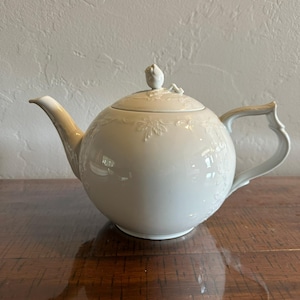 May include: A white porcelain teapot featuring a rounded body, a curved spout, and a handle with an angular design. The lid is topped with a sculpted rose finial, and the exterior surface is decorated with embossed floral and leaf patterns.