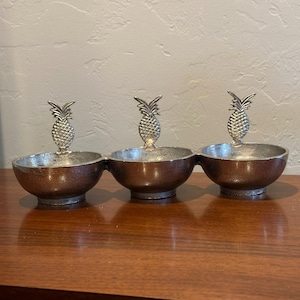 May include: A three-section serving dish with a silver pineapple handle on each section. The bowls are a dark bronze color, and the interior is silver. The dish is sitting on a wooden surface.