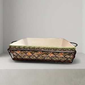 May include: A rectangular baking dish with a cream-colored interior and a decorative green and floral border. It sits in a dark metal wire basket with handles, suitable for serving. The basket has a floral pattern.