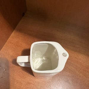 May include: A white ceramic mug with a square body and a rectangular handle. The mug has a small hole on the side near the top. The interior of the mug is white and the exterior is smooth.