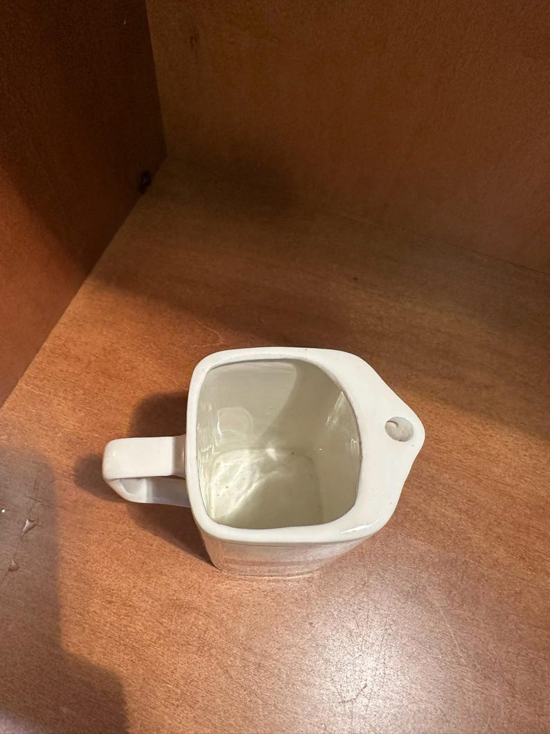 May include: A white ceramic mug with a square body and a rectangular handle. The mug has a small hole on the side near the top. The interior of the mug is white and the exterior is smooth.