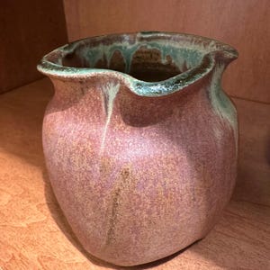 May include: A handcrafted ceramic vase with a unique, organic shape. The vase features a mottled, reddish-brown body with a green-glazed rim. The rim has a wavy, irregular edge, adding to its artistic appeal. The vase is displayed on a wooden surface.