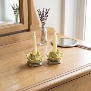 May include: Two yellow ceramic candle holders shaped like flowers, each holding a cream-colored candle. The candle holders sit on a wooden surface, with a small glass vase of lavender and a hand mirror in the background.