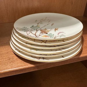 May include: A stack of six white ceramic plates with a gold rim. Each plate features a delicate design of autumn leaves in shades of green, orange, and brown. The plates are stacked on a wooden shelf.