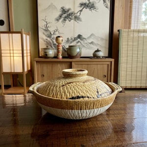 May include: A round, lidded ceramic cooking pot with two side handles. The exterior features a textured, tan-colored upper section with dark brown brushstroke accents and a smooth, white-glazed lower section with vertical ribbed detailing.