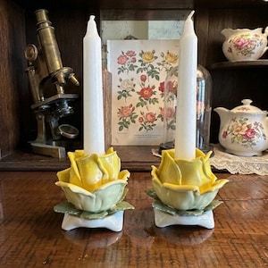 May include: A pair of ceramic candle holders shaped like blooming yellow roses with light green outer petals and white bases, each holding a tall, slender, unscented white taper candle. The rose petals are sculpted with realistic curves.
