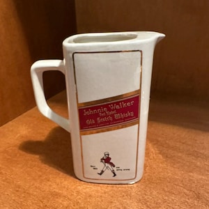 May include: White ceramic pitcher with a handle and a pouring spout. The front features a red and gold label with the text "Johnnie Walker Red Label Old Scotch Whisky" and a figure of a man walking. Gold trim accents the edges.