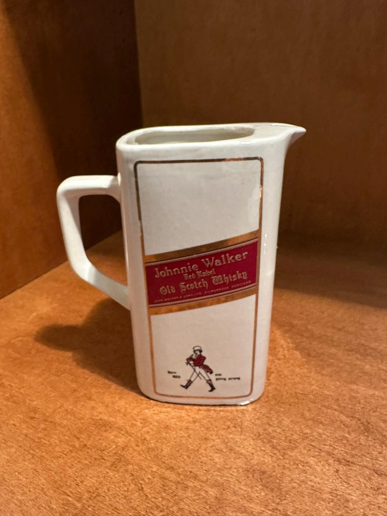 May include: White ceramic pitcher with a handle and a pouring spout. The front features a red and gold label with the text "Johnnie Walker Red Label Old Scotch Whisky" and a figure of a man walking. Gold trim accents the edges.
