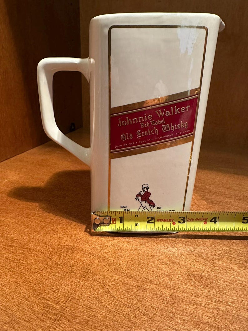 May include: A white ceramic pitcher with a rectangular shape and a handle. The pitcher features the "Johnnie Walker Red Label Old Scotch Whisky" logo in red and gold. The pitcher is approximately 4 inches wide.