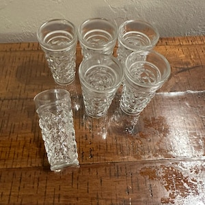 May include: A set of six clear glass tumblers featuring a textured diamond pattern on the exterior. Each tumbler has a smooth, circular rim and a cylindrical shape, with one glass positioned in the foreground and five arranged behind it.