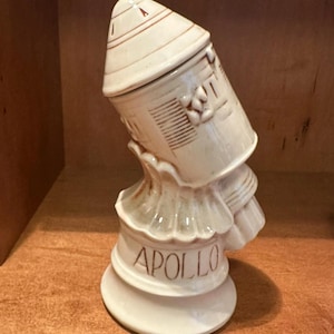 May include: Cream-colored ceramic Apollo space-themed figurine. The figurine features a rocket-shaped top with brown accents, a detailed building design, and the word "APOLLO" in brown lettering on the base. The figurine has a unique, tilted design.