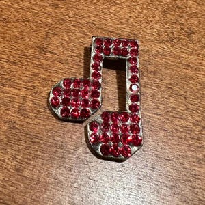 May include: A silver-toned musical eighth note, encrusted with numerous small, round, red rhinestones. The note is set against a brown wooden surface, creating a contrast between the shiny embellishments and the natural background.