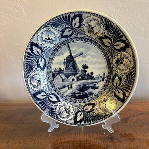 May include: A round ceramic plate featuring a dark blue and white transferware pattern. The center depicts a windmill in a rural landscape, while the wide border is decorated with intricate floral motifs and stylized leaves in a repeating design.