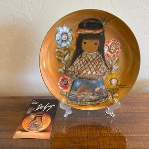 May include: A round decorative plate featuring a DeGrazia illustration of a child with long dark hair, a headband, and a patterned poncho, surrounded by colorful flowers on a golden-brown background. The plate rests on a clear stand next to a pamphlet.