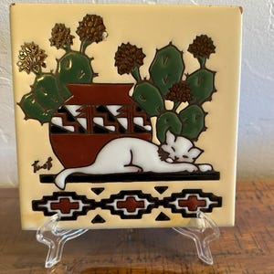 May include: Square ceramic tile with a cream-colored background. The tile features a white cat resting in front of a brown pot and green cacti. The tile is supported by a clear plastic stand. The tile has a Southwestern theme.