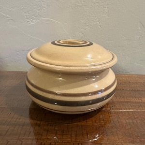 May include: A round, cream-colored ceramic lidded jar featuring horizontal bands in shades of brown and dark gray. The lid has a matching circular design with concentric rings, and the body of the jar has a smooth, glossy finish throughout.
