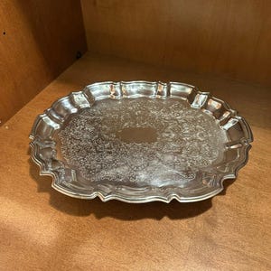 May include: A silver-colored, oval serving tray with ornate floral and scrollwork detailing. The tray has a scalloped edge and a reflective surface, suitable for serving food or displaying decorative items.