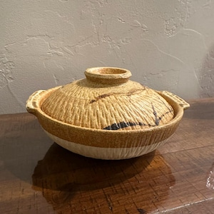 May include: A tan and cream-colored ceramic Japanese donabe pot with a lid. The pot has a textured, ridged surface with dark brown and black accents. The lid has a small handle and the pot has two side handles.
