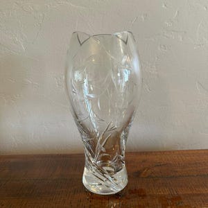 May include: A clear glass vase featuring an etched floral pattern with delicate leaves and stems. The vase has a tapered, bulbous body that narrows toward the base, which displays a distinct spiral or twisted design in the thick glass construction.