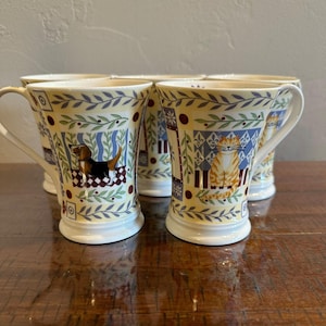May include: A set of ceramic mugs featuring a pale yellow background with a decorative border of green leaves and purple accents. Each mug displays a distinct animal illustration, including a brown and black dog and an orange tabby cat.