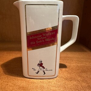 May include: White ceramic pitcher with a rectangular design featuring the "Johnnie Walker Red Label Old Scotch Whisky" logo in red and gold. Includes a drawing of a man walking and the text "Born 1820" and "still going strong."
