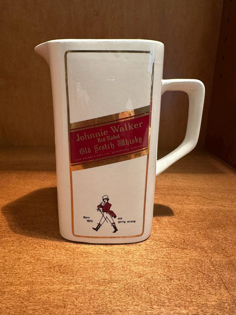 May include: White ceramic pitcher with a rectangular design featuring the "Johnnie Walker Red Label Old Scotch Whisky" logo in red and gold. Includes a drawing of a man walking and the text "Born 1820" and "still going strong."