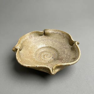 May include: A handmade, beige ceramic bowl with a unique, wavy rim design. The bowl has a shallow, circular interior with a spiral pattern at the base. The exterior has a textured, rustic appearance, suggesting a natural, earthy aesthetic.