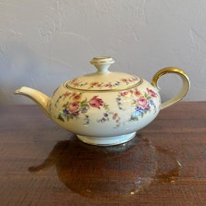 May include: A cream-colored porcelain teapot featuring a delicate floral pattern of pink roses, blue forget-me-nots, and green foliage. The teapot includes a matching lid with a small knob, a curved spout, and a gold-trimmed handle and rim.