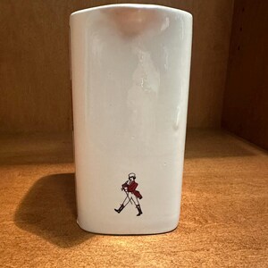 May include: A white ceramic pitcher with a rectangular shape. The pitcher features a small illustration of a man in a red coat and black boots. The pitcher is sitting on a wooden surface.