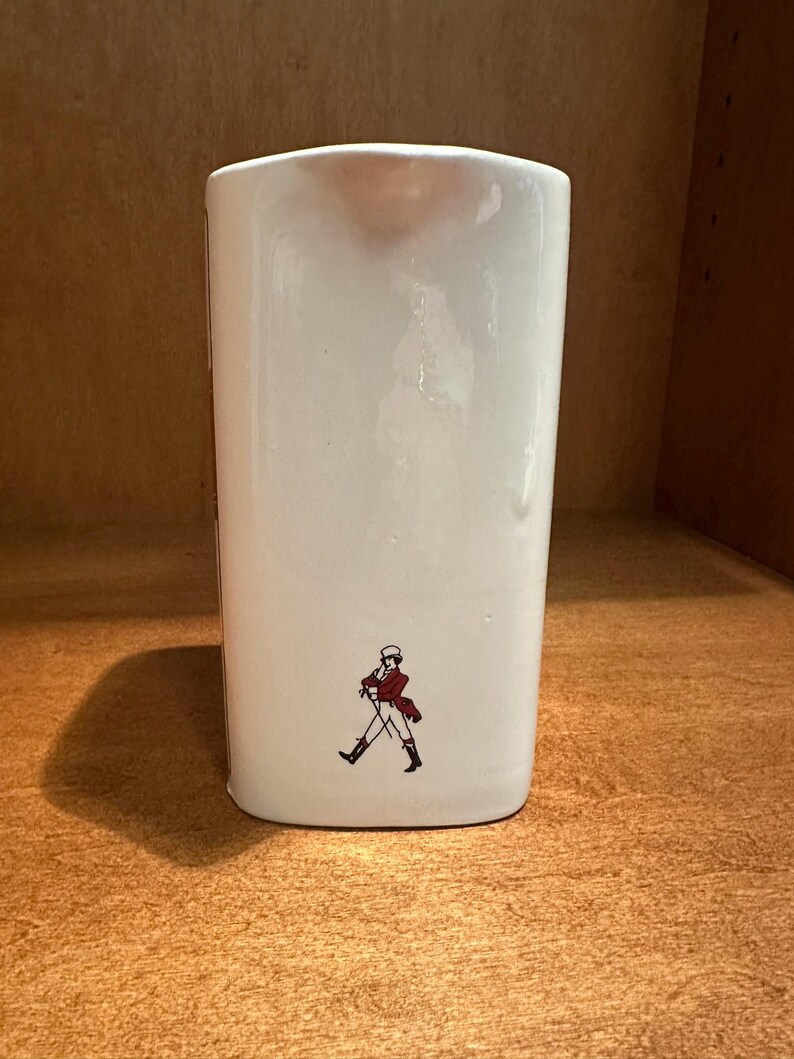 May include: A white ceramic pitcher with a rectangular shape. The pitcher features a small illustration of a man in a red coat and black boots. The pitcher is sitting on a wooden surface.