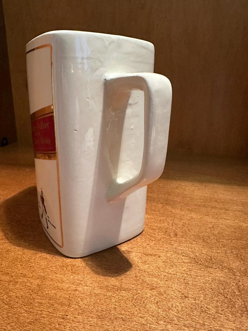 May include: White ceramic mug with a rectangular shape and a large handle. The mug features a red and gold label with text and a figure. The mug is sitting on a wooden surface.