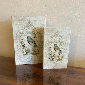 May include: Two decorative boxes with a vintage design. Each box features a detailed illustration of a bird perched on a branch with a nest, set against a backdrop of script and ornate scrollwork. The boxes are rectangular and vary in size.
