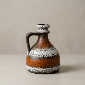 May include: A ceramic pitcher with a handle, featuring horizontal bands of white, orange, and brown. The top has a textured, speckled design. The pitcher is a decorative item.
