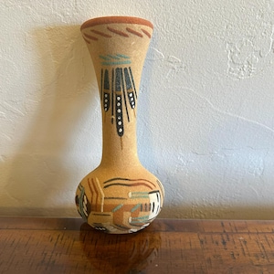 May include: A tan-colored vase with a narrow neck and rounded base. The vase features hand-painted geometric designs in shades of brown, black, blue, and white. The rim is a darker brown color. The vase is approximately 8 inches tall.