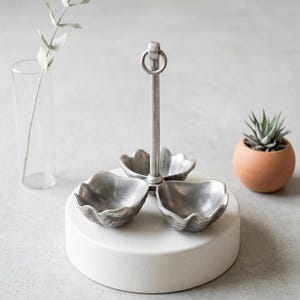May include: A three-sectioned, silver-toned serving dish with a central handle and ring, set on a white, circular base. The dish has a seashell-like design. A small potted succulent and a clear vase with a sprig of greenery are in the background.