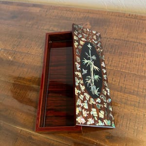May include: Rectangular wooden box with a hinged lid. The lid is decorated with an oval black inlay featuring bamboo and butterflies. The box's exterior is adorned with iridescent shell pieces, and the interior is a dark reddish-brown.