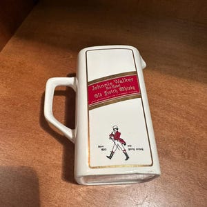 May include: White ceramic pitcher with a handle, featuring the "Johnnie Walker Red Label Old Scotch Whisky" logo in red and gold. The pitcher also has an illustration of the Johnnie Walker striding man and the text "Born 1820, still going strong."