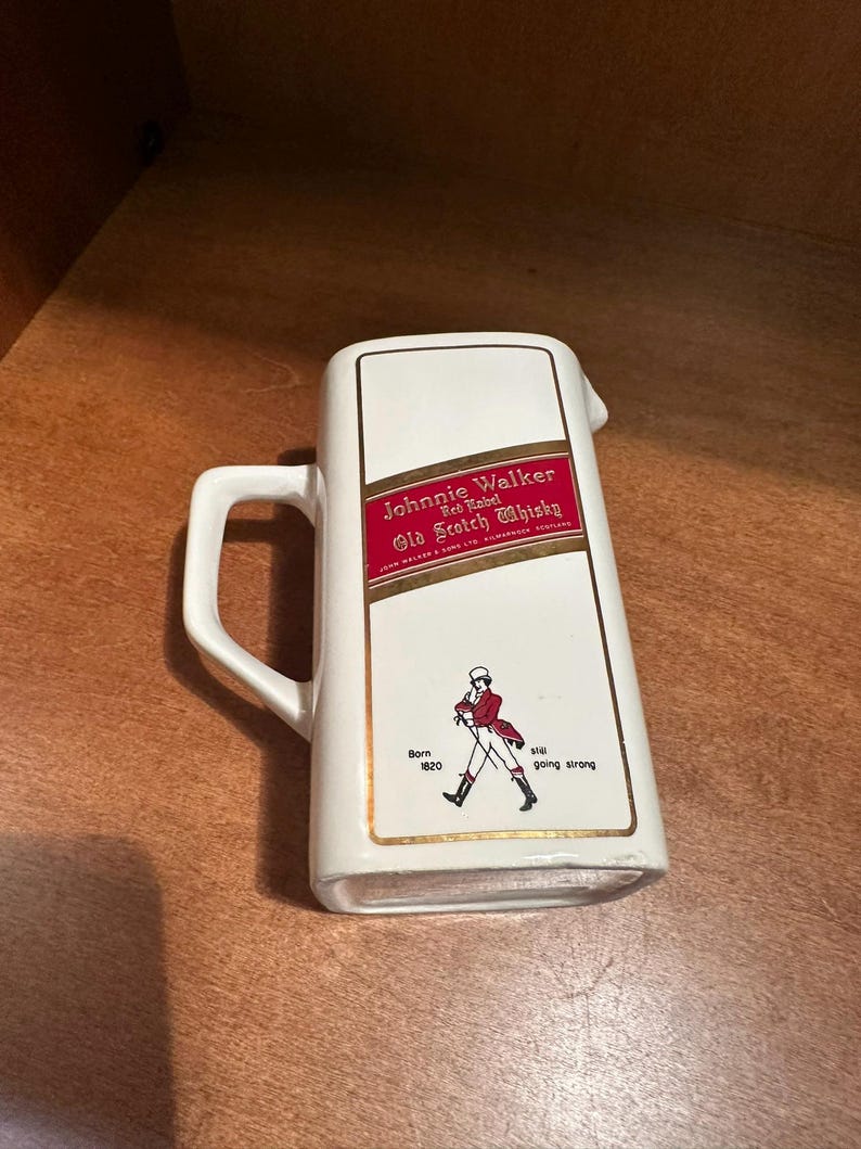 May include: White ceramic pitcher with a handle, featuring the "Johnnie Walker Red Label Old Scotch Whisky" logo in red and gold. The pitcher also has an illustration of the Johnnie Walker striding man and the text "Born 1820, still going strong."