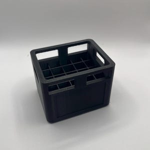 Könnte beinhalten: Eine kleine, schwarze, rechteckige Kiste mit einem Gitter aus Trennwänden im Inneren. Die Kiste hat rechteckige Öffnungen an den Seiten und oben für eine einfache Handhabung. Die Kiste wird wahrscheinlich zum Organisieren oder Aufbewahren kleiner Gegenstände verwendet.