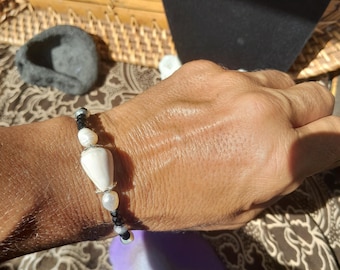 Könnte beinhalten: Ein handgefertigtes Armband mit einer großen, weiß geformten Muschelperle, umgeben von kleineren weißen Perlen und schwarzen Perlen. Das Armband ist an einem schwarzen Band aufgereiht und wird an einem Handgelenk gezeigt.
