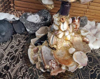 Könnte beinhalten: Ein dekorativer Ständer mit einem runden Fuß und einer Mittelsäule, die dicht mit verschiedenen weißen, beigen und braunen Muscheln in goldfarbenem Klebematerial besetzt ist und ein muschelverziertes, rundes Oberteil trägt.