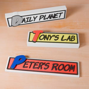 May include: Three white rectangular signs with black borders and colorful letters. The top sign says "DAILY PLANET", the middle sign says "TONY'S LAB", and the bottom sign says "PETER'S ROOM".