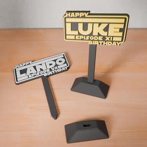 May include: Two black stands with a white and yellow sign on each. The sign on the left says "Happy Lando Episode VII Birthday". The sign on the right says "Happy Luke Episode XI Birthday".