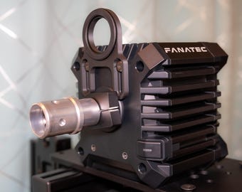 May include: A black Fanatec racing wheel base with a silver metal shaft and a circular handle. The base has a ribbed design and the brand name "FANATEC" is visible on the top.