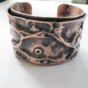 May include: A copper cuff bracelet with a textured, hammered finish. The bracelet has a small, round, gold-colored accent in the center.