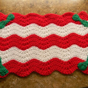 Puede incluir: Una alfombrilla rectangular de ganchillo con un patrón de ondas en zigzag y rayas horizontales alternas en hilo rojo brillante y blanco. Las cuatro esquinas de la alfombrilla están decoradas con un pequeño lazo de ganchillo verde.